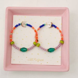 May include: A pair of colorful beaded hoop earrings with green, blue, orange, and pink beads. The earrings are displayed on a pink tray with the text "Jill Makes" on a white card.
