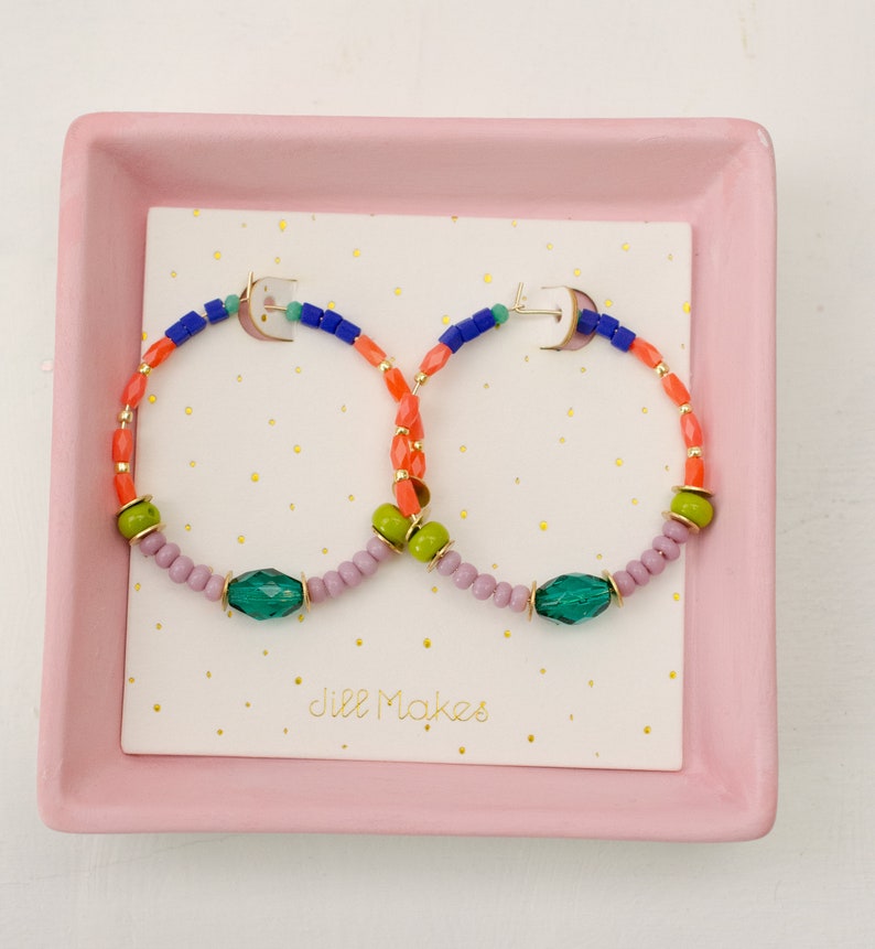 May include: A pair of colorful beaded hoop earrings with green, blue, orange, and pink beads. The earrings are displayed on a pink tray with the text "Jill Makes" on a white card.