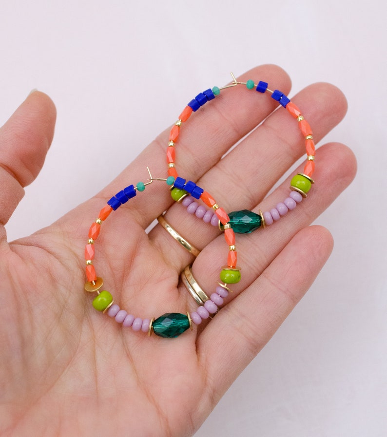 May include: A pair of hoop earrings with a multi-colored beaded design. The earrings feature orange, green, blue, and purple beads, as well as a larger green gemstone bead.