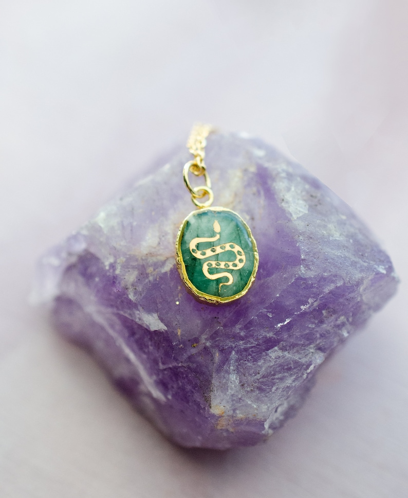 May include: A gold chain necklace with a green oval pendant featuring a gold snake design. The pendant is resting on a purple amethyst crystal.