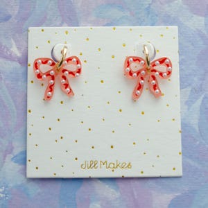 May include: A pair of bow-shaped earrings with a red and white design, adorned with small white pearls. The earrings are attached to gold-colored hoops and displayed on a white card with gold dots and the text "Jill Makes".
