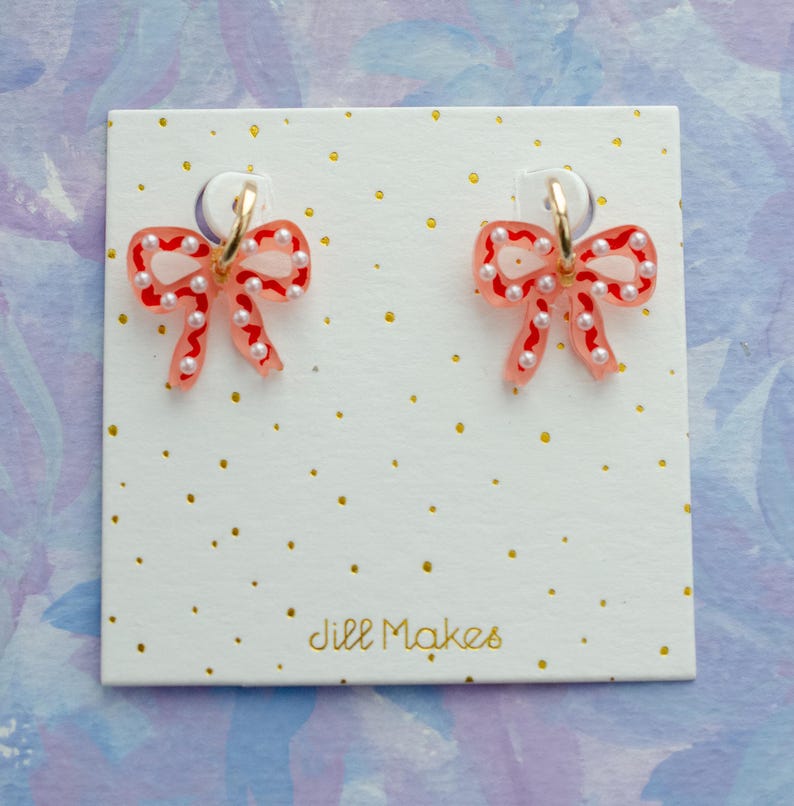May include: A pair of bow-shaped earrings with a red and white design, adorned with small white pearls. The earrings are attached to gold-colored hoops and displayed on a white card with gold dots and the text "Jill Makes".