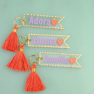 May include: Three clear acrylic keychains with gold accents and red tassels. Each keychain features a white polka dot background with a strawberry graphic and a name: Adora, Emma, and Camilla.
