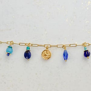 May include: Gold chain bracelet with blue and turquoise glass beads and charms, including a star, a crescent moon, a zodiac symbol, and a tag that says "Joy Always".