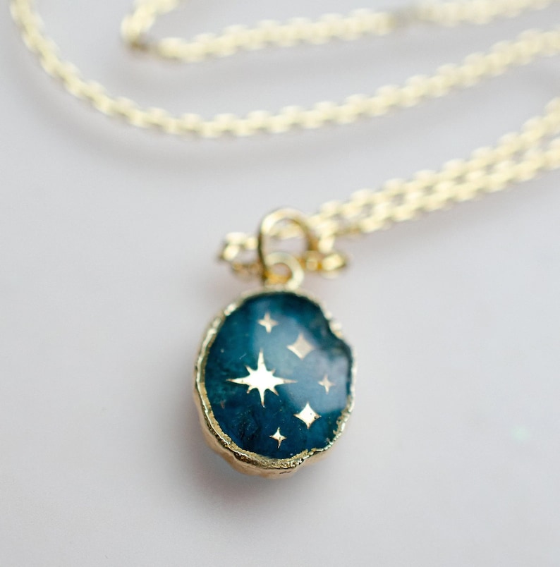 Op de afbeelding: Een gouden ketting met een donkerblauwe hanger met een gouden ster en kleinere gouden sterren op een donkerblauwe achtergrond.