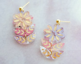 Könnte beinhalten: Ein Paar Hängeohrringe mit floralem Design. Die Ohrringe sind aus klarem Acryl mit rosa und violetten Akzenten und goldenen Details gefertigt. Die Blumen sind übereinander gelegt und haben einen zarten, femininen Look.