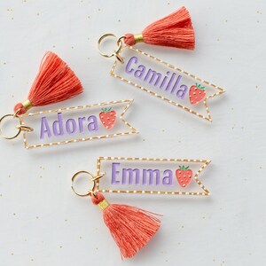 May include: Three personalized keychains with a pink and white strawberry design. Each keychain has a gold ring, a pink tassel, and a clear acrylic tag with a white dotted border. The tags are personalized with the names "Adora", "Camilla", and "Emma" in purple lettering.