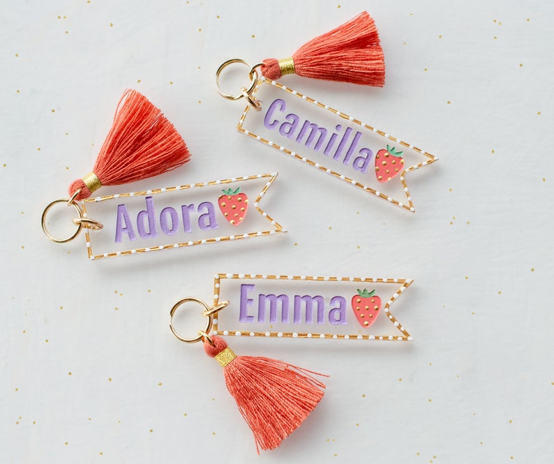 May include: Three personalized keychains with a pink and white strawberry design. Each keychain has a gold ring, a pink tassel, and a clear acrylic tag with a white dotted border. The tags are personalized with the names "Adora", "Camilla", and "Emma" in purple lettering.