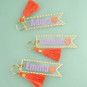 May include: Three clear acrylic keychains with gold trim and a pink tassel. Each keychain has a different name, Adora, Camilla, and Emma, written in purple with a strawberry graphic.