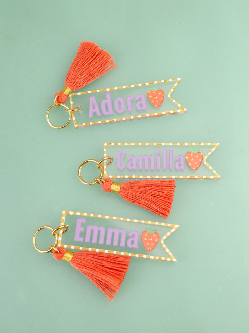 May include: Three clear acrylic keychains with gold trim and a pink tassel. Each keychain has a different name, Adora, Camilla, and Emma, written in purple with a strawberry graphic.