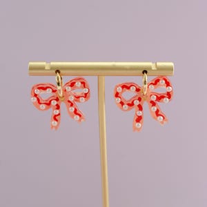 May include: A pair of bow-shaped earrings with a red outline and filled with a translucent pink hue. The earrings are adorned with small, white pearl-like beads and gold-colored hoops. The earrings are displayed on a gold-colored earring stand.