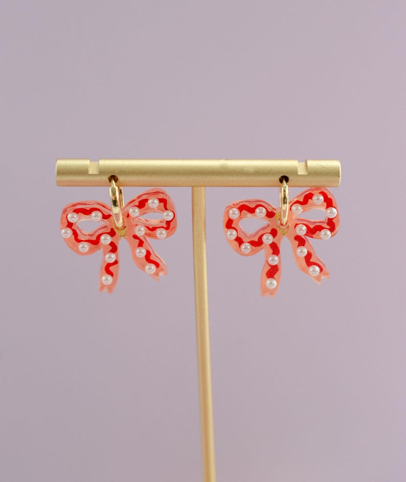 May include: A pair of bow-shaped earrings with a red outline and filled with a translucent pink hue. The earrings are adorned with small, white pearl-like beads and gold-colored hoops. The earrings are displayed on a gold-colored earring stand.