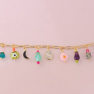 May include: A gold charm bracelet with a variety of colorful charms, including a crescent moon, a flower, a small bottle, and a heart. The bracelet has a small tag that says "Jill Makes".
