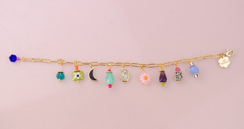 May include: A gold charm bracelet with a variety of colorful charms, including a crescent moon, a flower, a small bottle, and a heart. The bracelet has a small tag that says "Jill Makes".