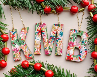 May include: Four wooden ornaments shaped like the letters K, M, B, and a smaller letter B, each painted with a pink, green, and white floral pattern. The ornaments are hanging from gold chains against a light pink background.
