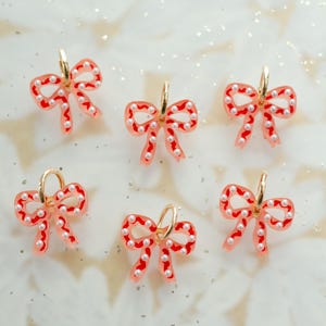 May include: Six bow-shaped earrings with gold hoops. The bows are a translucent pink with red trim and small white pearl accents. The earrings are arranged on a white surface with glitter.