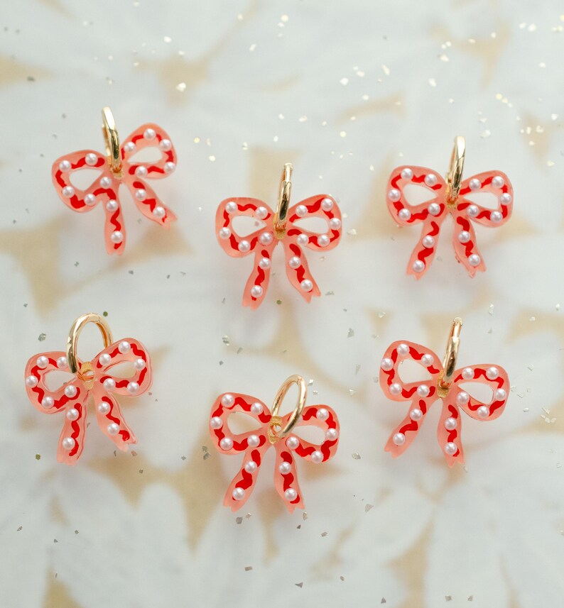 May include: Six bow-shaped earrings with gold hoops. The bows are a translucent pink with red trim and small white pearl accents. The earrings are arranged on a white surface with glitter.