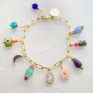 May include: A gold chain bracelet with multiple charms, including a flower, a crescent moon, a gemstone, a letter J, and a daisy. The charms are colorful and have a variety of textures.