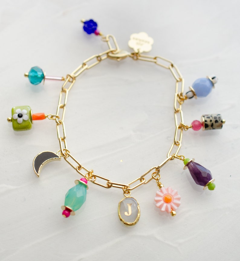 May include: A gold chain bracelet with multiple charms, including a flower, a crescent moon, a gemstone, a letter J, and a daisy. The charms are colorful and have a variety of textures.