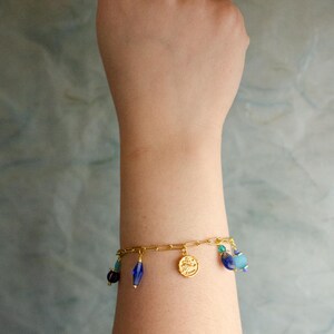 May include: A gold chain bracelet with blue and turquoise glass beads and a gold charm. The bracelet is on a person's wrist.
