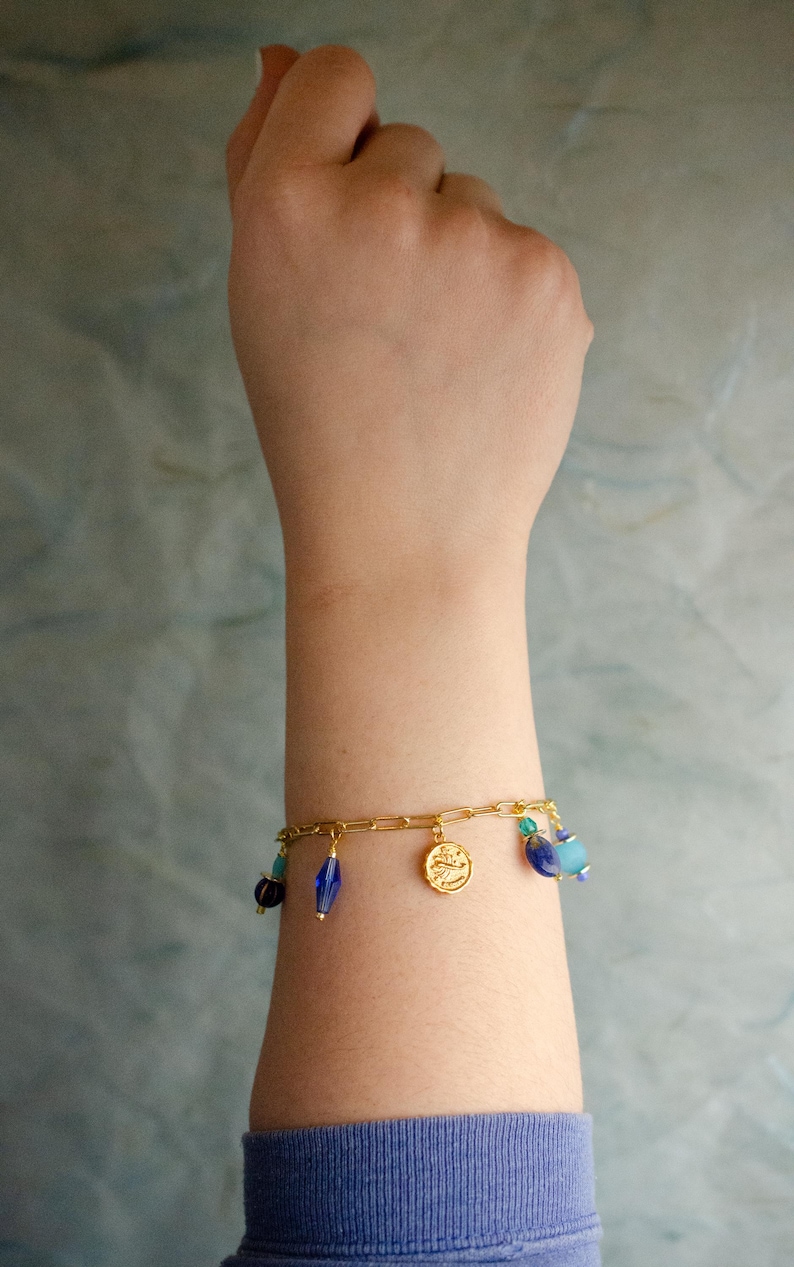 May include: A gold chain bracelet with blue and turquoise glass beads and a gold charm. The bracelet is on a person's wrist.