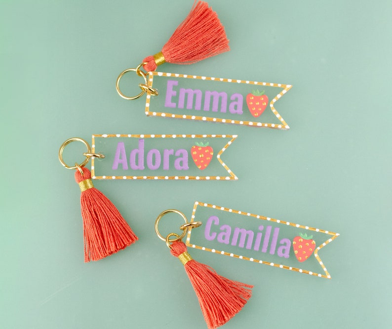 May include: Three acrylic keychains with gold tassels. Each keychain has a white background with gold polka dots and a pink strawberry graphic. The keychains are personalized with the names "Emma", "Adora", and "Camilla".