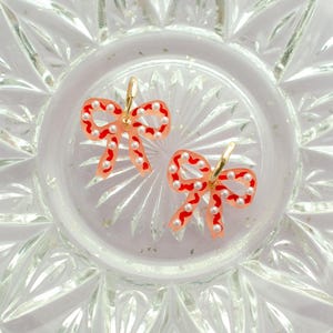 May include: A pair of bow-shaped earrings. The earrings feature a translucent peach-colored base with red outlines and are adorned with small, white pearl-like beads. The earrings have gold-colored hoops.