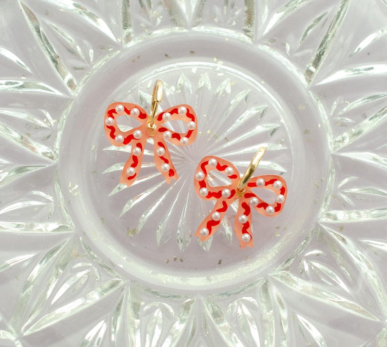 May include: A pair of bow-shaped earrings. The earrings feature a translucent peach-colored base with red outlines and are adorned with small, white pearl-like beads. The earrings have gold-colored hoops.