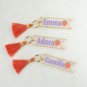 May include: Three clear acrylic keychains with gold trim and a pink tassel. Each keychain has a white name, "Emma", "Adora", and "Camilla", with a pink strawberry graphic.