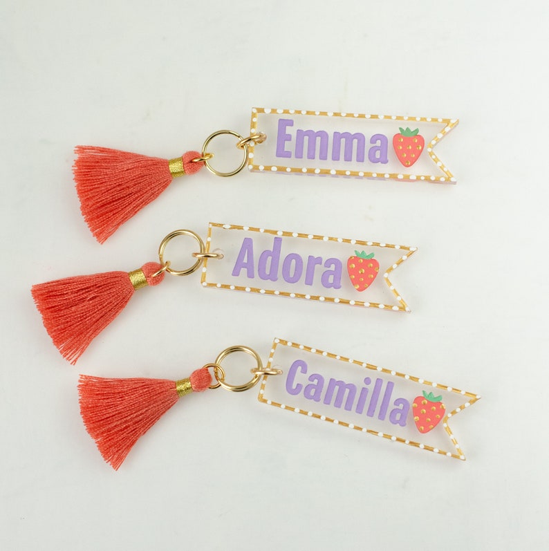 May include: Three clear acrylic keychains with gold trim and a pink tassel. Each keychain has a white name, "Emma", "Adora", and "Camilla", with a pink strawberry graphic.