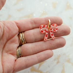 May include: A clear, bow-shaped earring with red outlines and white pearl accents. The earring has a gold-colored hoop. The bow design is a festive accessory.