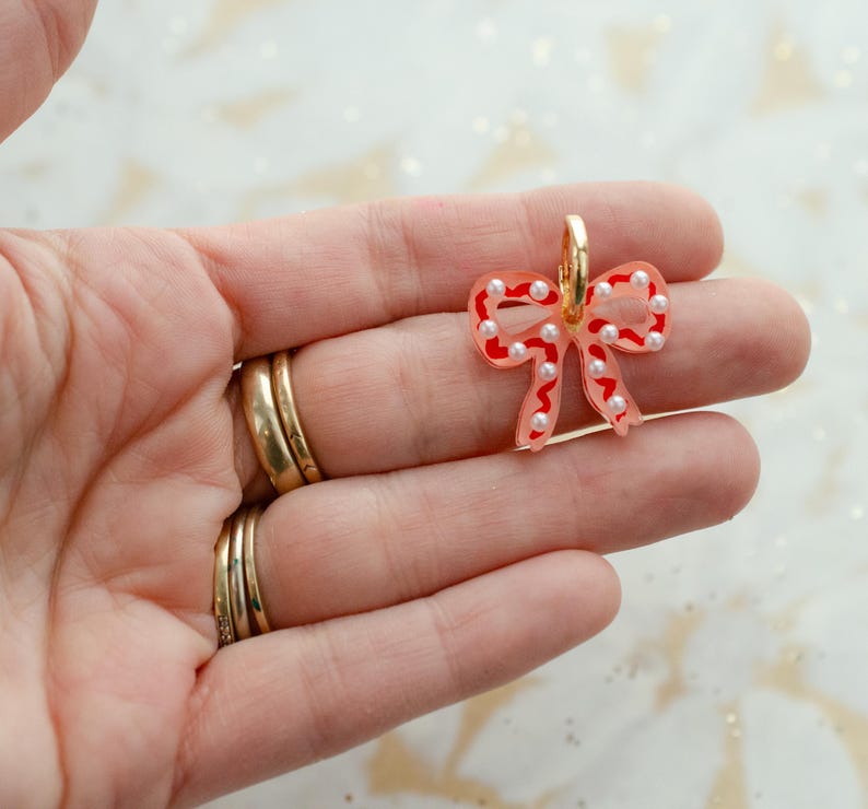 May include: A clear, bow-shaped earring with red outlines and white pearl accents. The earring has a gold-colored hoop. The bow design is a festive accessory.