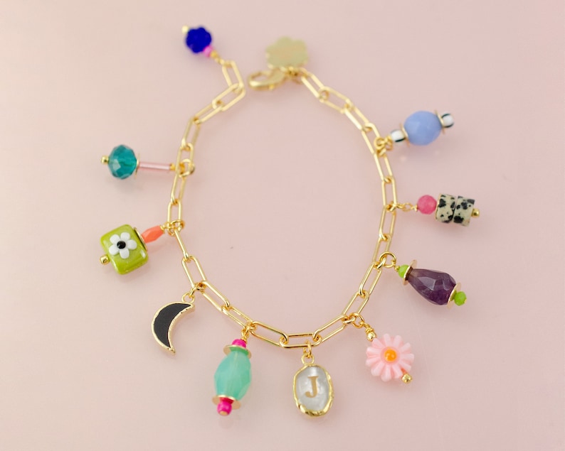 May include: A gold chain bracelet with multiple charms, including a crescent moon, a flower, a letter J, and various colored stones.