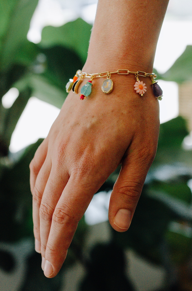 May include: A gold chain bracelet with colorful charms, including a flower, a letter J, and a crescent moon.