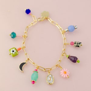 May include: A gold chain bracelet with multiple charms, including a crescent moon, a flower, a green square with a white flower, a light blue oval, a purple teardrop, a light blue round bead, a pink flower, and a clear oval with the letter "J" inside.