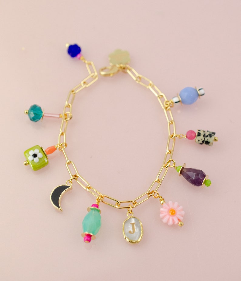 May include: A gold chain bracelet with multiple charms, including a crescent moon, a flower, a green square with a white flower, a light blue oval, a purple teardrop, a light blue round bead, a pink flower, and a clear oval with the letter "J" inside.