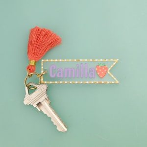 May include: A pink and gold key chain with a clear acrylic tag that says "Camilla" and a small strawberry graphic. The key chain has a coral tassel and a silver key.
