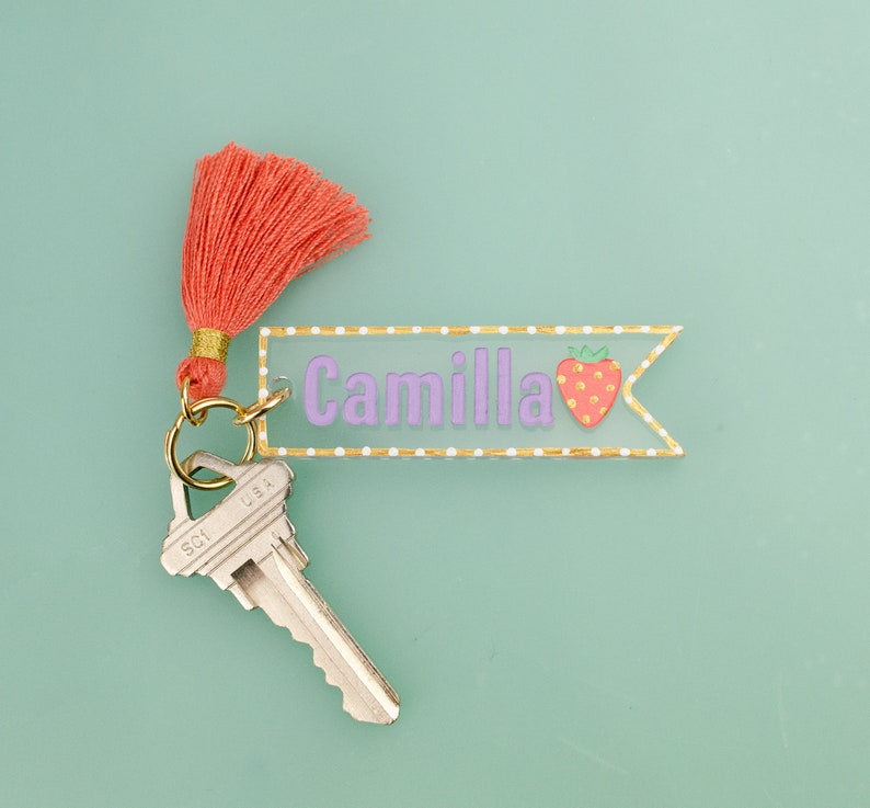 May include: A pink and gold key chain with a clear acrylic tag that says "Camilla" and a small strawberry graphic. The key chain has a coral tassel and a silver key.