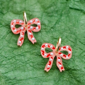 May include: A pair of bow-shaped earrings. The earrings are light pink with red accents and small white pearl-like beads. The earring hoops are gold-colored. The earrings are on a green textured surface.