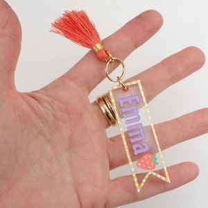 May include: A clear acrylic keychain with a gold border and white polka dots. The keychain features the name "Emma" in purple lettering and a red and green strawberry illustration. A coral tassel hangs from the top of the keychain.