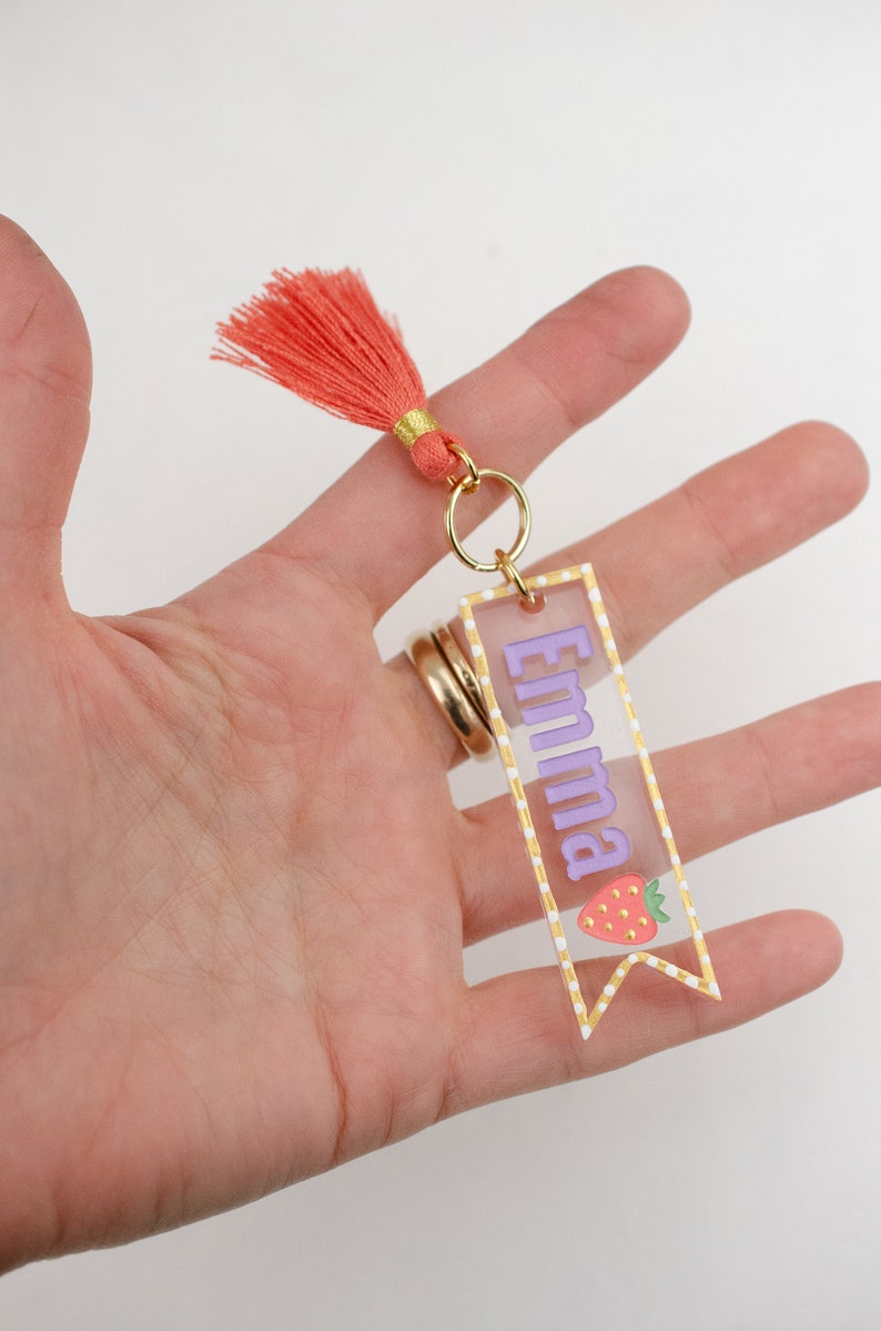 May include: A clear acrylic keychain with a gold border and white polka dots. The keychain features the name "Emma" in purple lettering and a red and green strawberry illustration. A coral tassel hangs from the top of the keychain.
