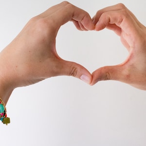 May include: A person's hands forming a heart shape, with a gold charm bracelet on their wrist. The bracelet features colorful beads, flowers, and a charm with the letter "S".