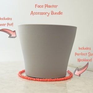 Op de afbeelding: Grijze gezichtsplantenbak met een koraalkleurige kralenketting rond de basis. De tekst "Includes Inner Pot!" staat boven de plantenbak en "Includes Perfect Size Necklace!" staat onder de plantenbak.