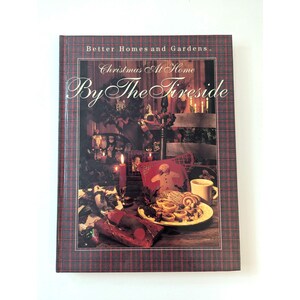 May include: A red and black plaid book cover with the title "Christmas At Home By The Fireside" and a photo of a cozy Christmas scene with a fireplace, decorations, and baked goods.