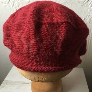 May include: A red knitted beret on a mannequin head. The beret is made of a soft, textured yarn.