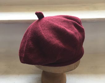 May include: A burgundy knit beret with a small, rounded top detail. The hat is displayed on a wooden hat form against a white background. The beret is made of a textured knit material.