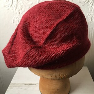 May include: A deep red knitted beret, sitting on a wooden hat form.