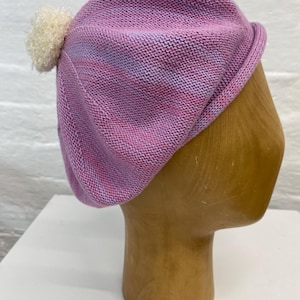 May include: A pink knit beret with a white pom-pom. The beret is displayed on a wooden head form. The knit fabric has a mix of pink and purple hues.