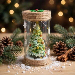 Pode incluir: Um frasco cilíndrico de vidro transparente contendo uma árvore de Natal em miniatura verde texturizada com glitter dourado e pequenos ornamentos vermelhos. O frasco apresenta um acabamento em corda de juta no topo e na base.