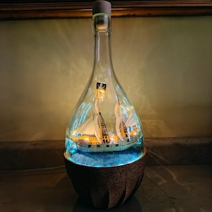 Può includere: Una bottiglia di vetro trasparente contenente un modellino di nave con vele bianche e una bandiera pirata nera. La nave poggia su una base di materiale blu all'interno della bottiglia, illuminata da luci calde. La bottiglia ha un tappo di sughero.